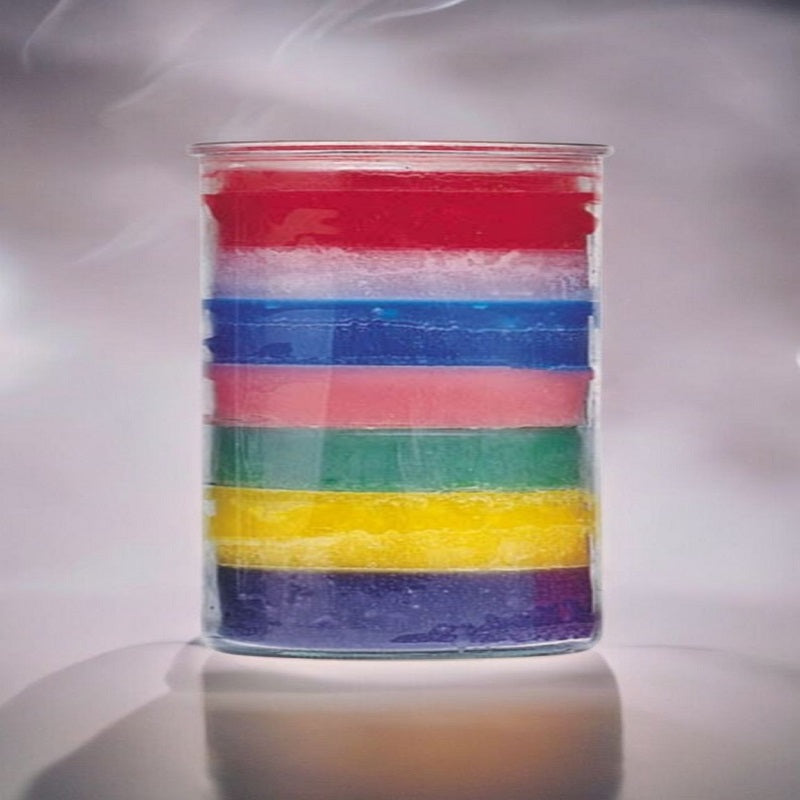 A tall 7-layer unscented candle on a gray glass table. Dim teal light accentuates vibrant wax colors and creates a calming, spiritual atmosphere.